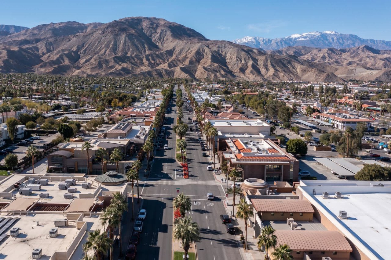 Aerial view of a palm-lined desert city street with rugged mountains in the distance.