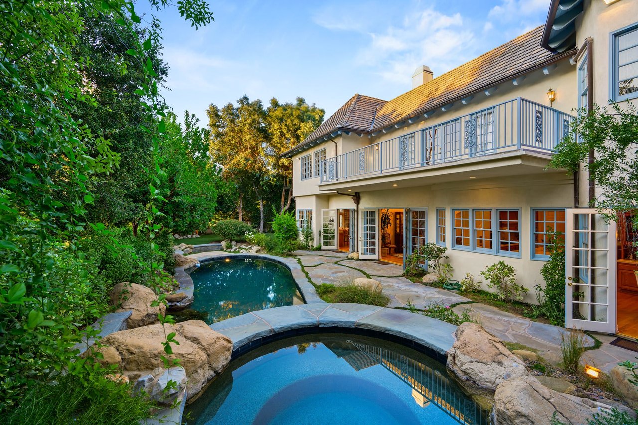 A luxurious two-story house with a light-colored exterior and a blue-railed balcony overlooking a landscaped backyard.