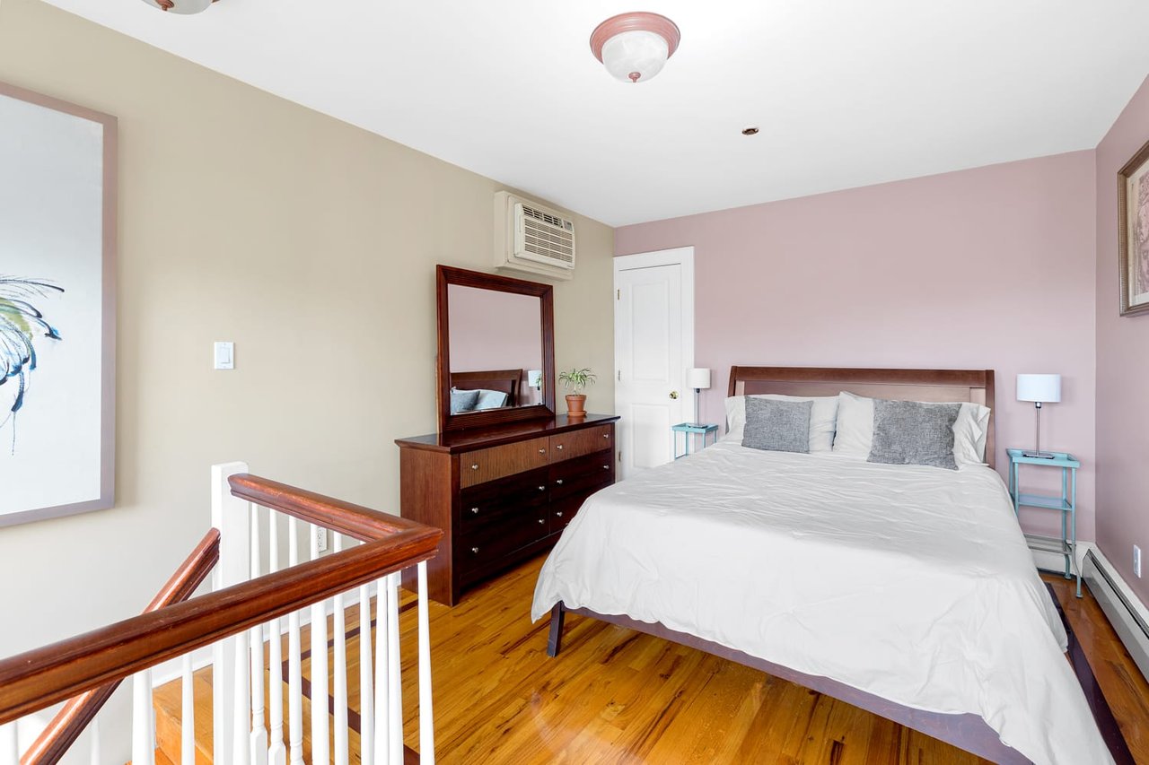 Bedroom with a large white-covered bed, dark wood dresser, pink wall, and hardwood floors.