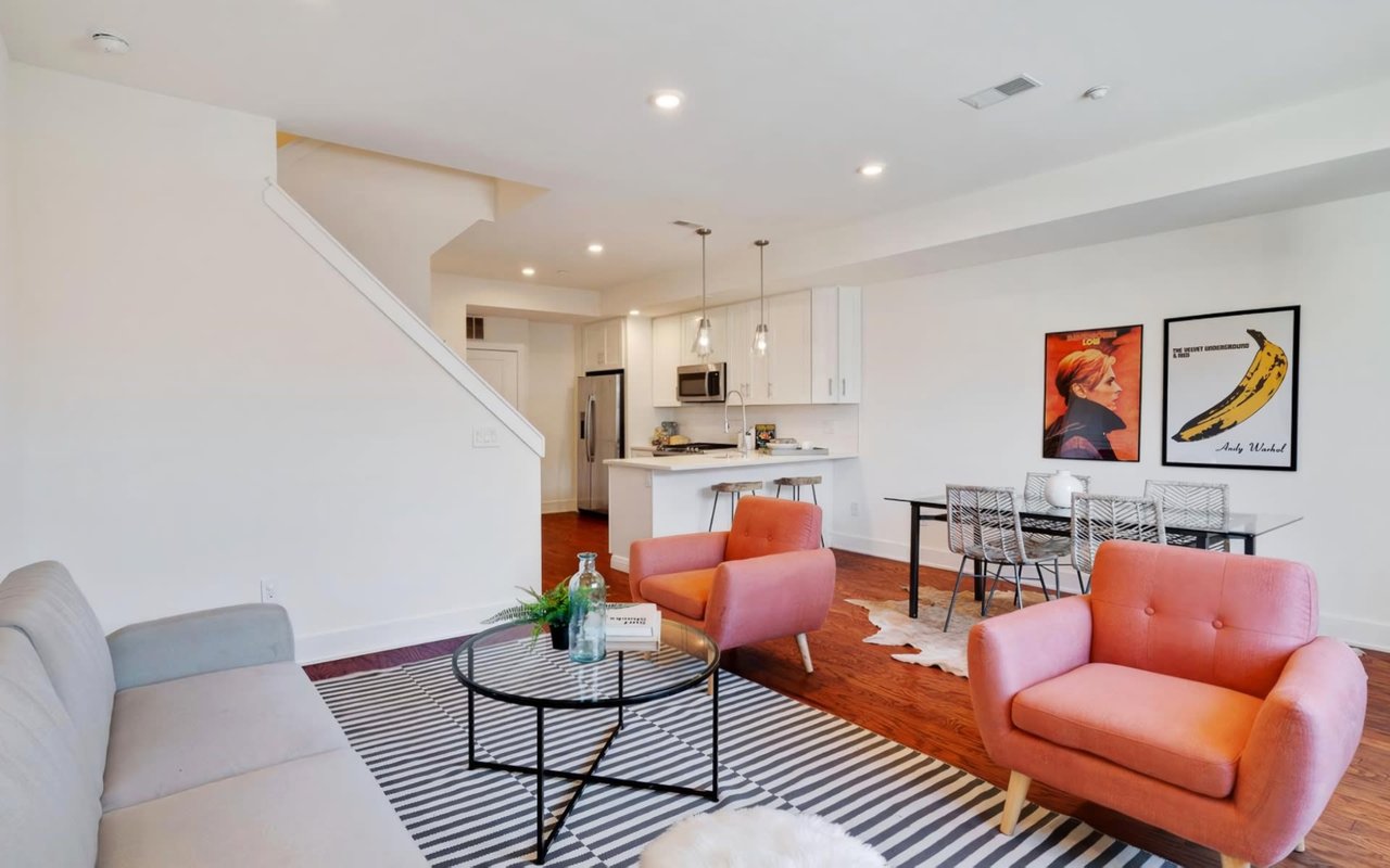 Gray sectional, pink armchairs, glass coffee table, dining area, white kitchen.