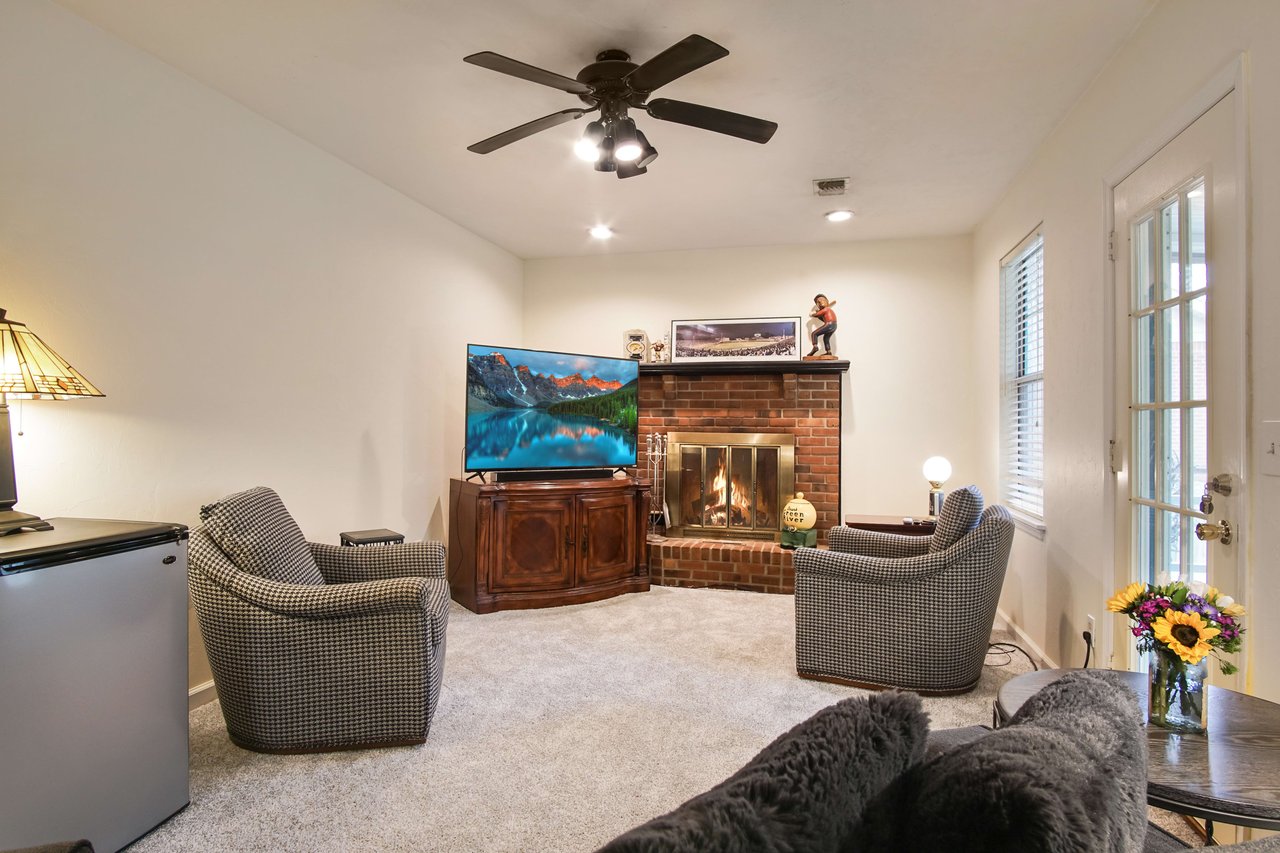 A cozy living room with comfortable seating, including chairs and a sofa. The room features a fireplace, a ceiling fan, and large windows allowing natural light to enter.