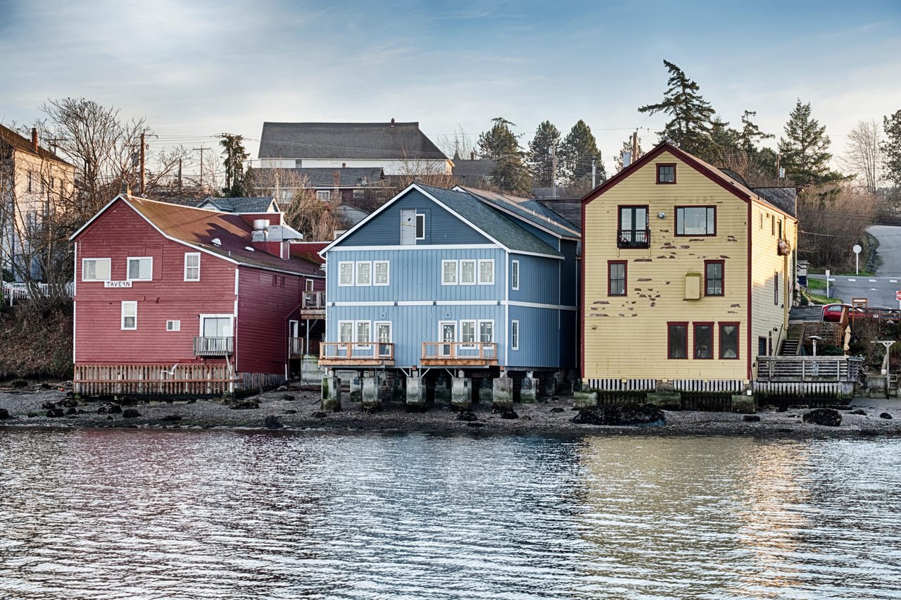 Row of houses on stilts next to a body of water. The houses are painted white and have green roofs.