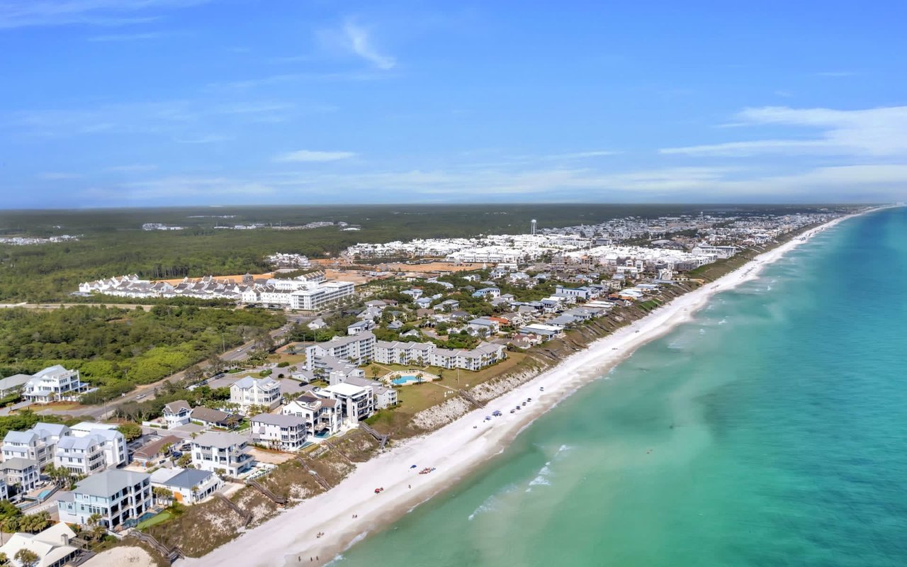 An aerial view of a coastal town with white houses, a sandy beach, and clear blue water.