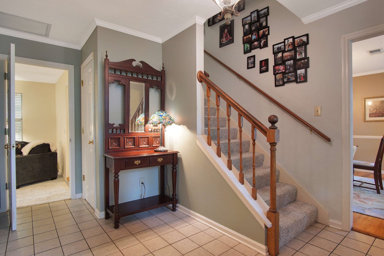 A view of a hallway with a staircase leading to the upper floor. The hallway is decorated with pictures on the wall, and there is a small table with decorative items.