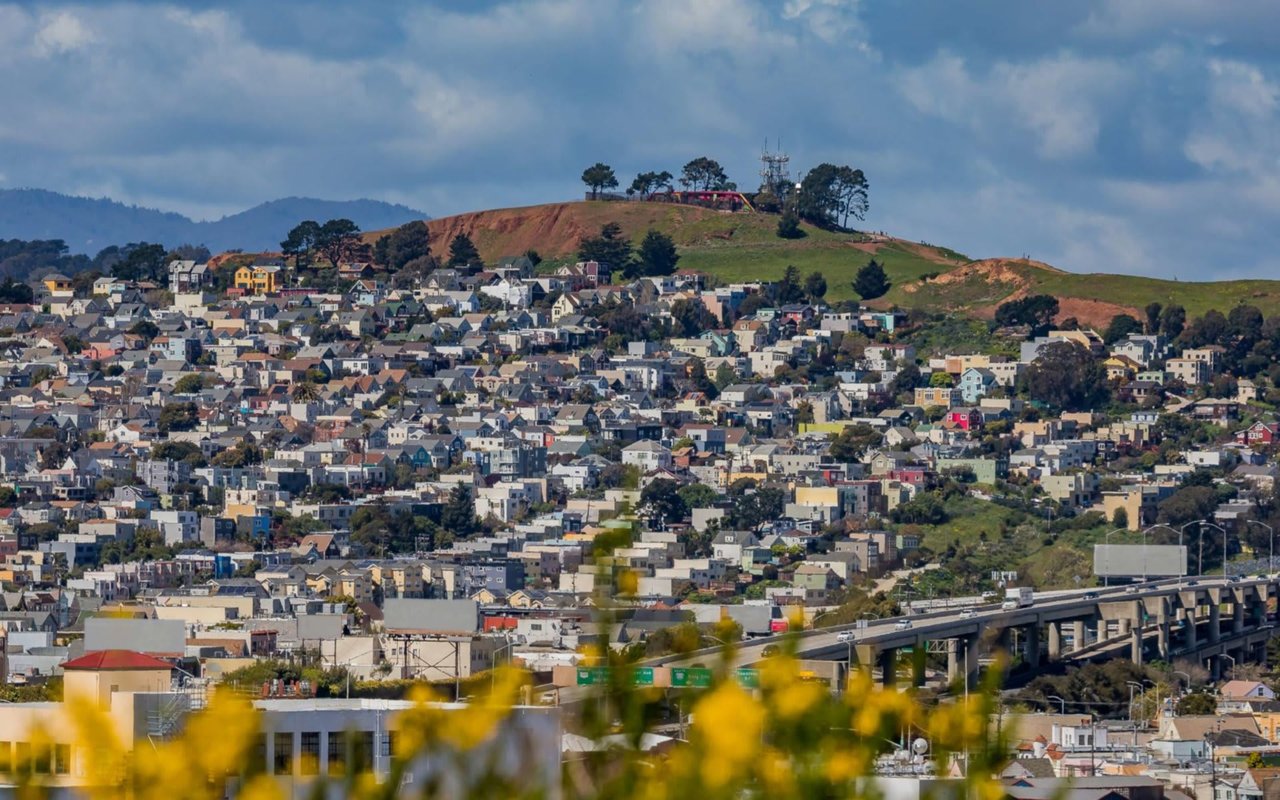 A scenic view of Bernal Heights, San Francisco, featuring densely packed homes of various colors and styles cascading down the hillside. The top of the hill is sparsely vegetated with some trees and communication towers. In the foreground, yellow flowers and a highway interchange are visible. The scene is set under a cloudy blue sky.