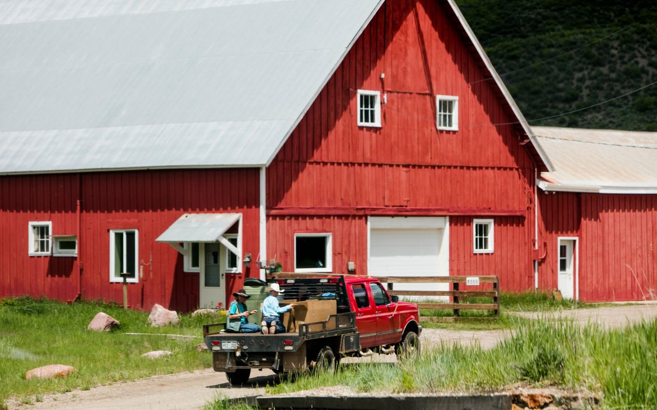 Historic red barn in Old Snowmass with a pickup truck and farm workers in the foreground, showcasing the rural agricultural heritage that continues in this scenic Colorado valley surrounded by rolling green hills.