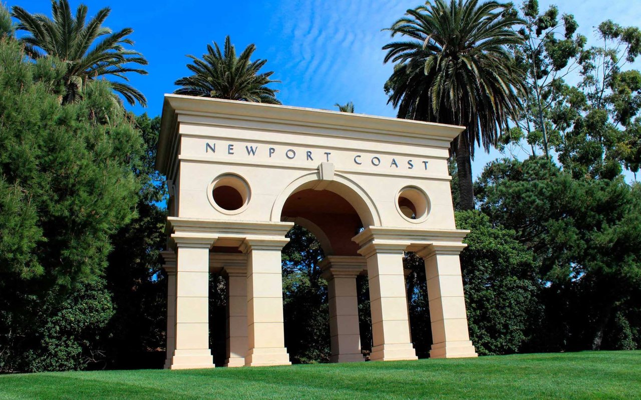 A white arched doorway of Newport Coast surrounded by palm trees and lush green bushes