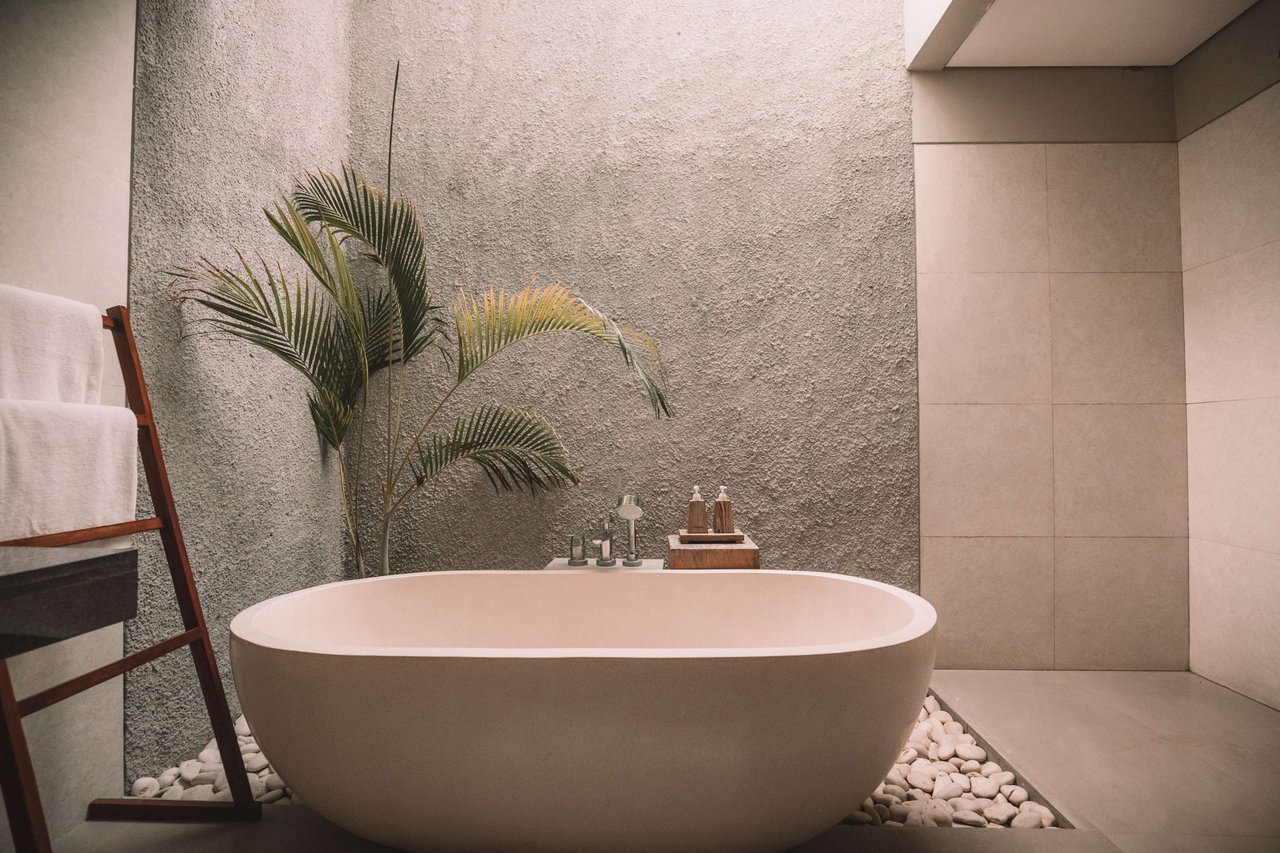 A modern bathroom with a freestanding oval bathtub, a palm tree, and a ladder with towels.