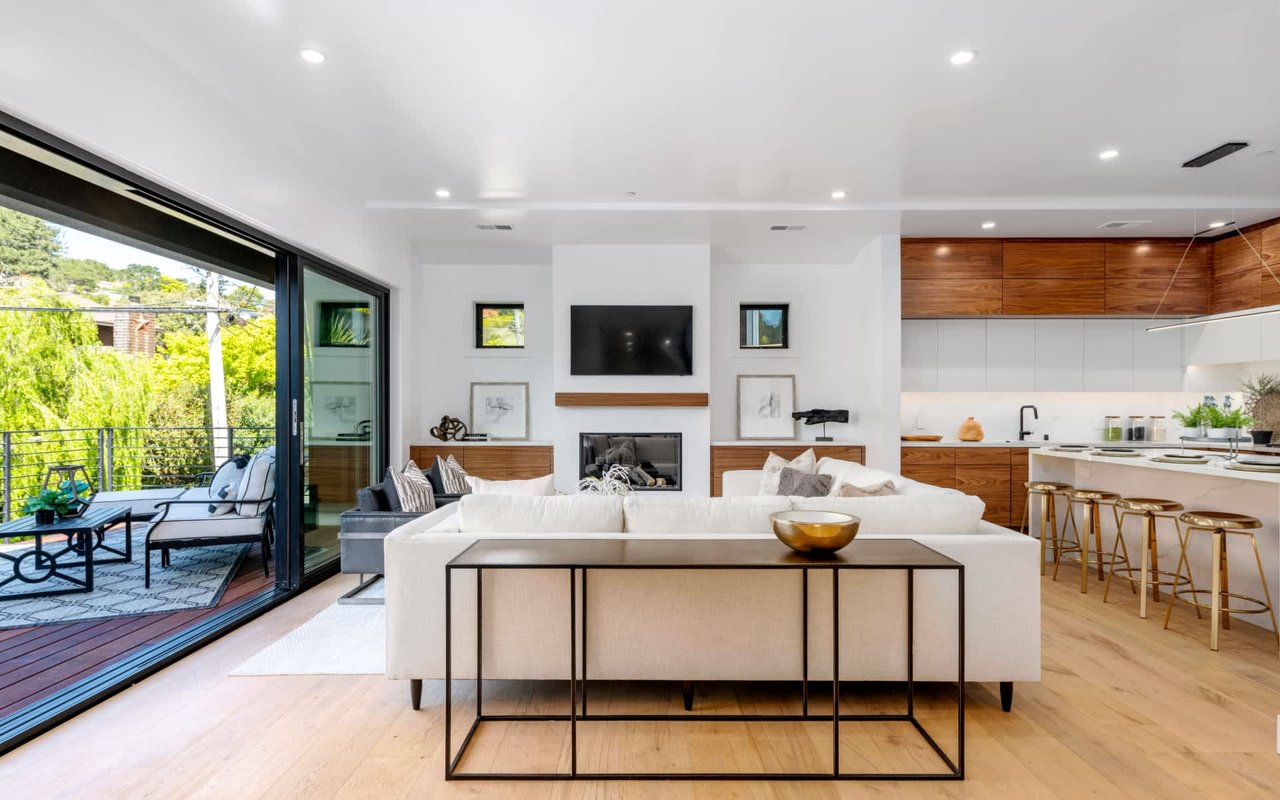Open-plan living room and kitchen with a white sectional, fireplace, wood cabinetry, and doors to a leafy deck.