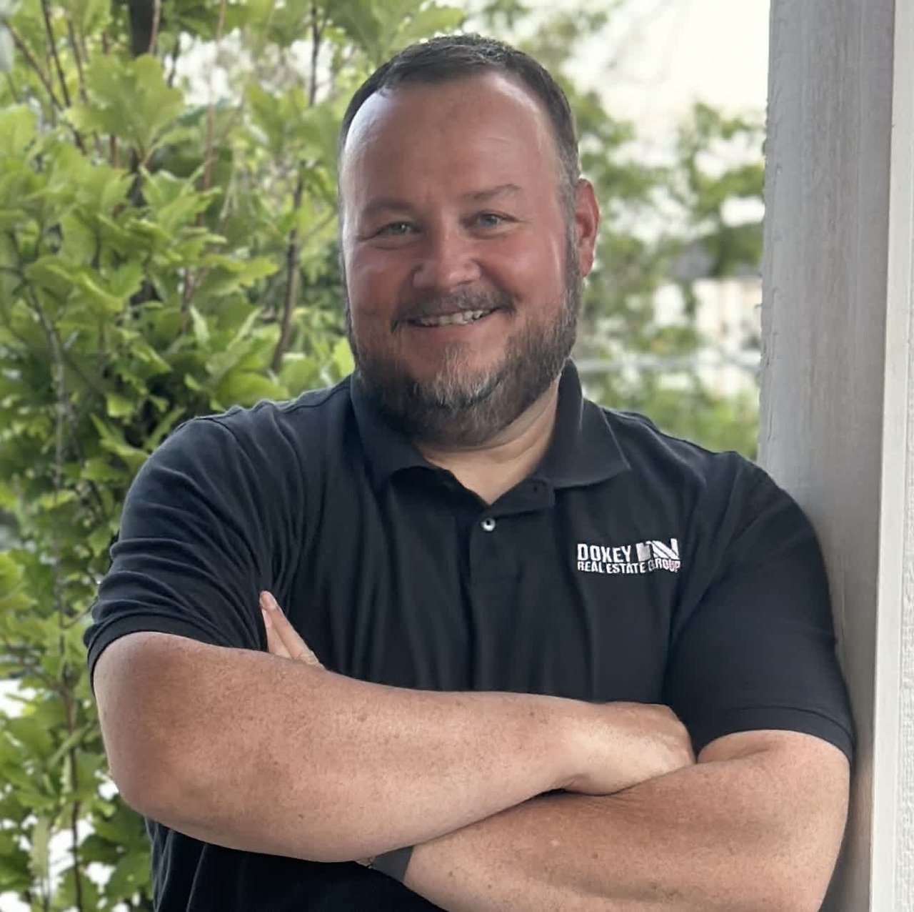 A bearded man wearing a black polo shirt with the "Doxey Real Estate Group" logo on the chest. He is standing outdoors, smiling with his arms crossed in a relaxed posture. Green foliage and a white column are visible in the background.