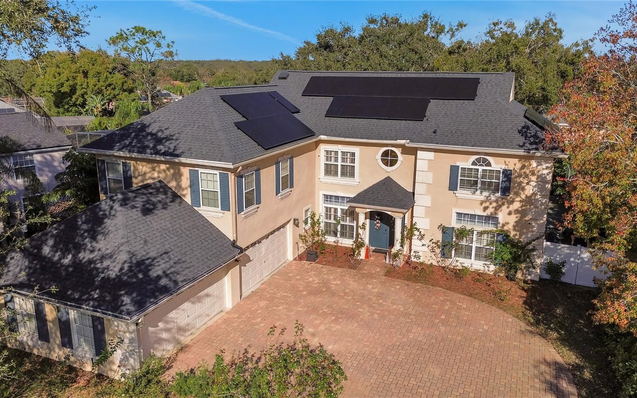 A two-story house with solar panels on its roof, a curved driveway to an attached two-car garage, surrounded by trees.