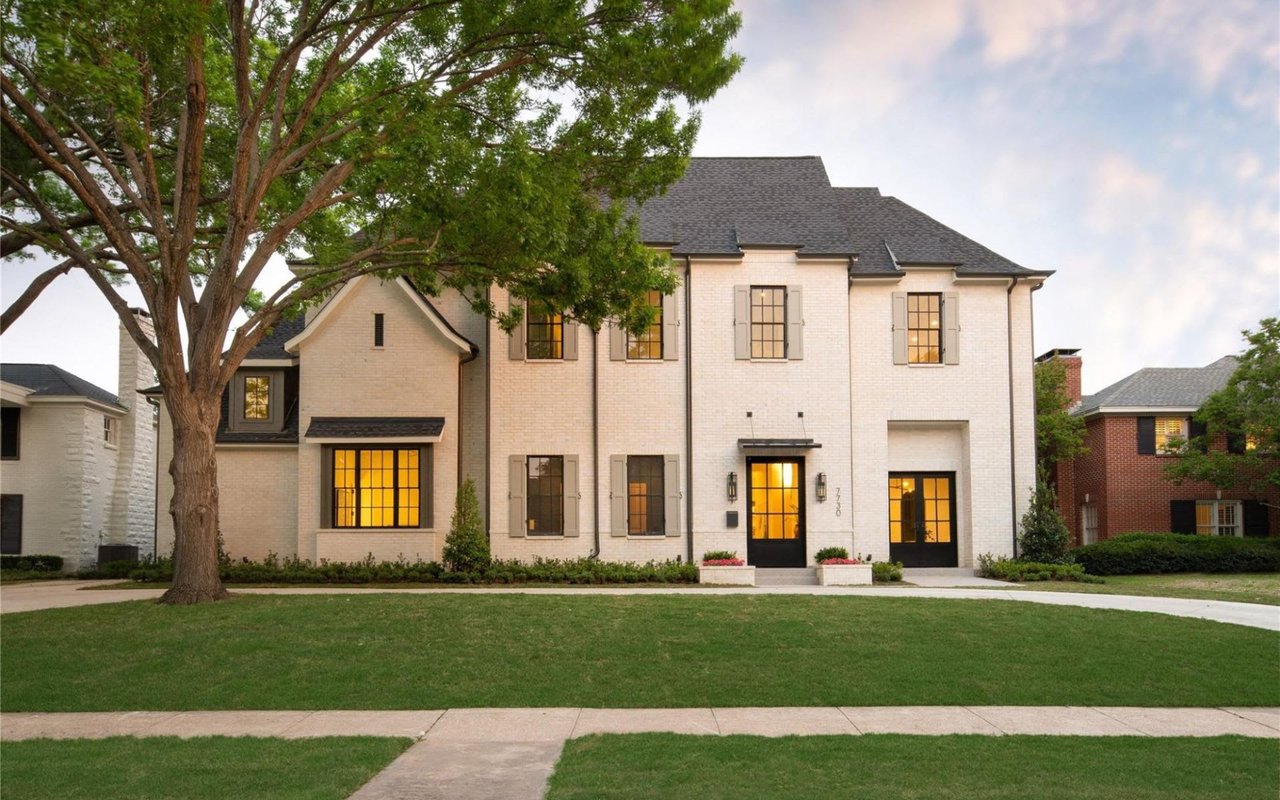 A two-story white house with a covered porch, a front lawn, a paved street, and a sidewalk.