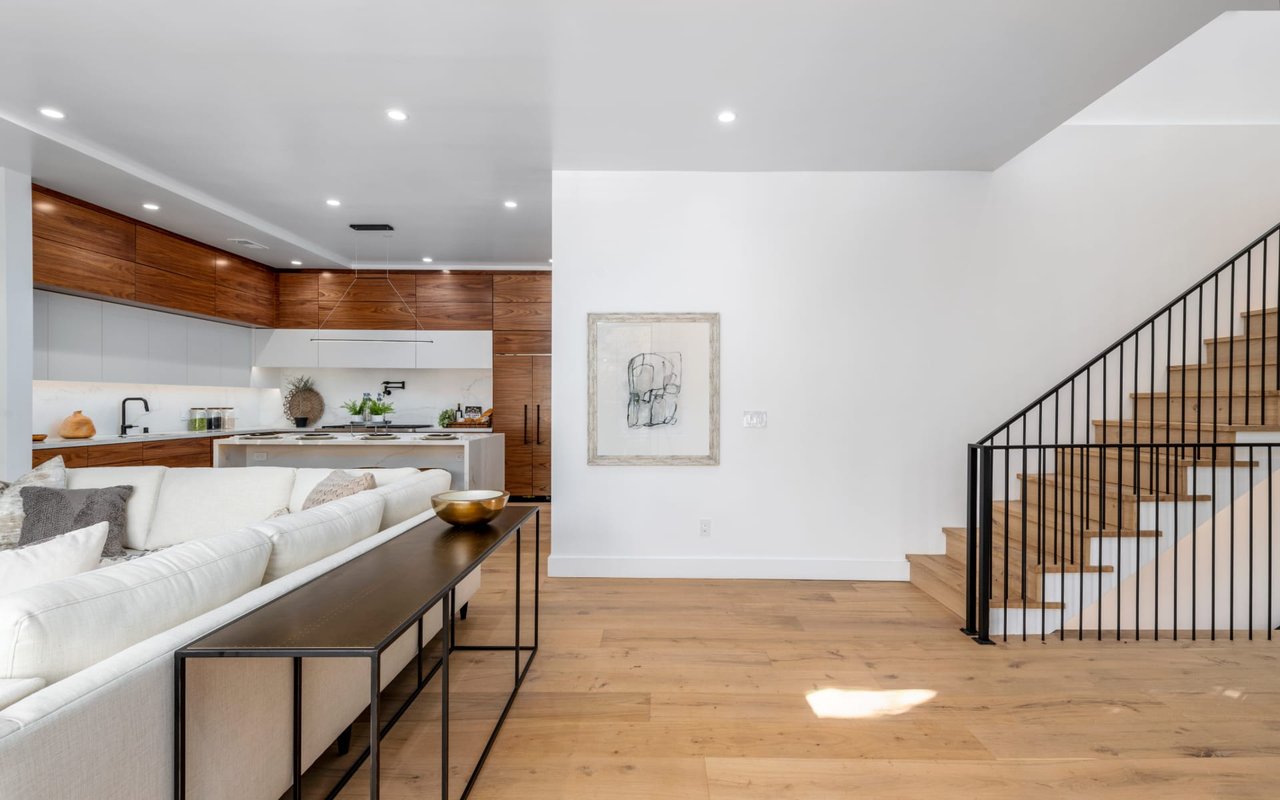 Open-plan living room with a white sectional, wood-cabinet kitchen, and staircase with black railing.