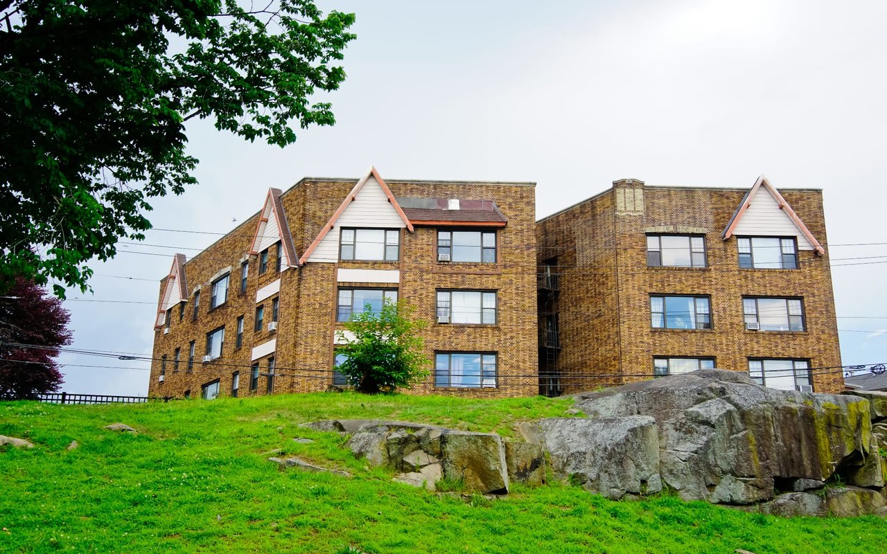 A large, red brick apartment building on top of a grassy hill, surrounded by a few scattered trees.