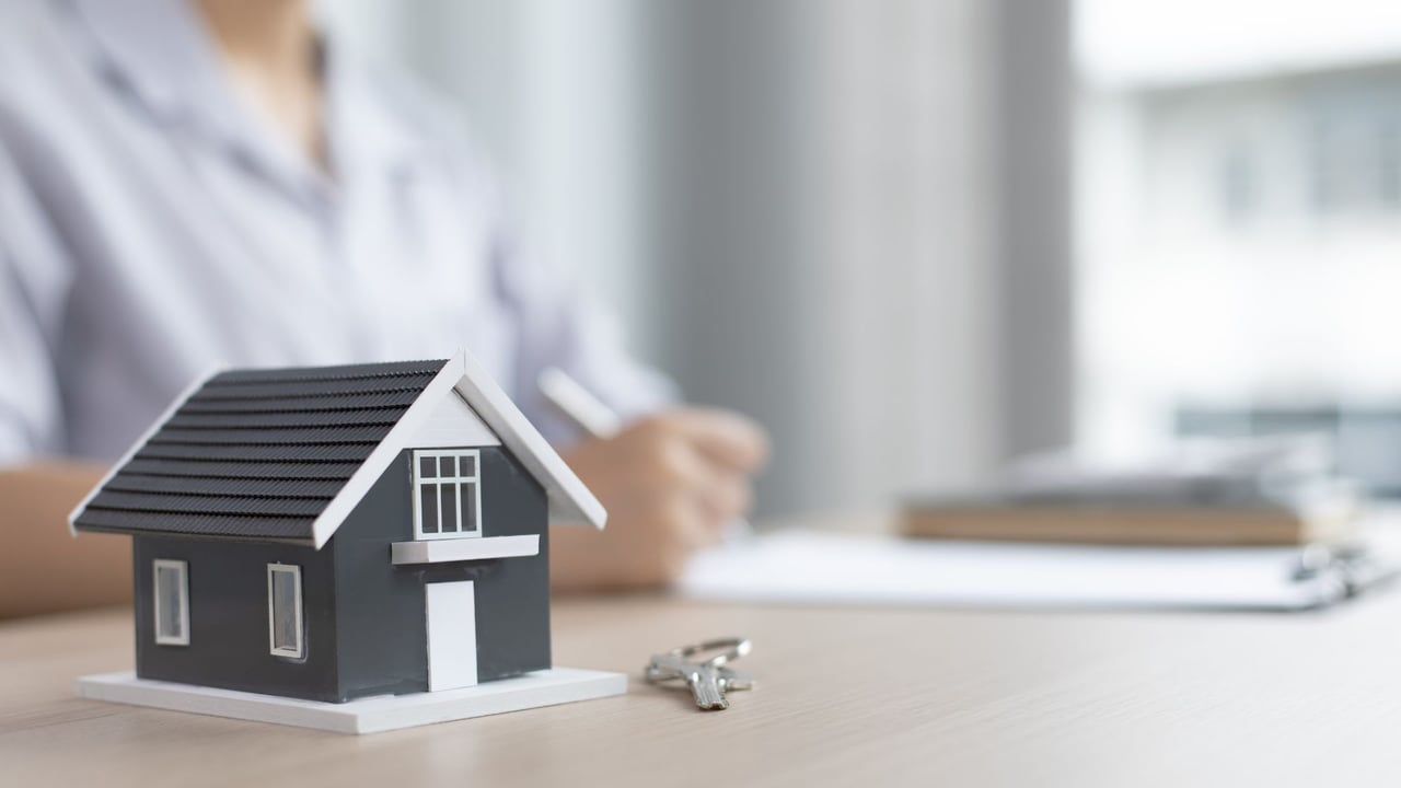 A miniature house and keys sit beside paperwork as a person writes in the background.