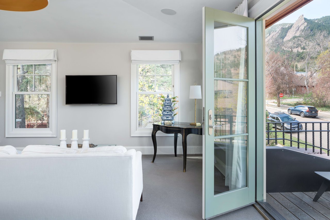 White living room with TV, balcony doors, and Flatirons mountain view. Features white sofa and blue ceramic decor.