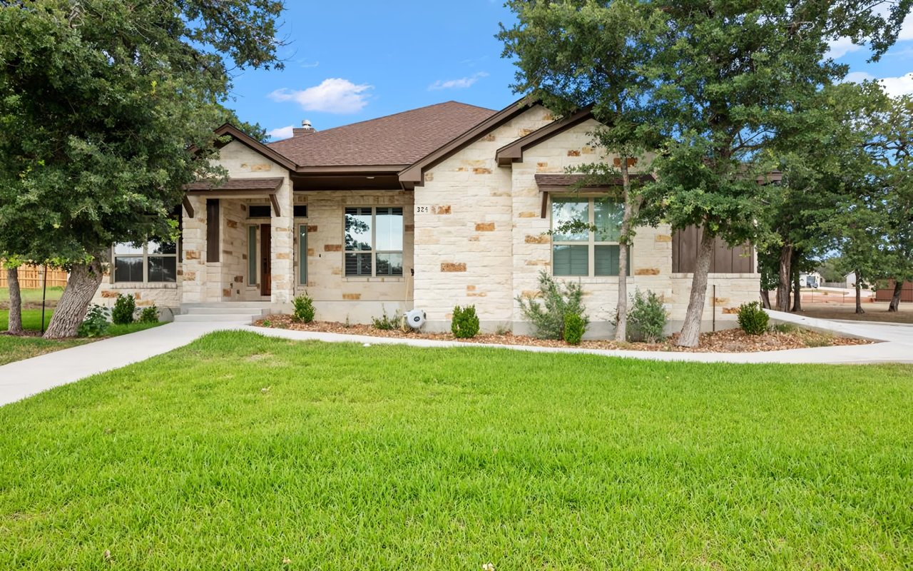 A large, single-story house with light-colored stone siding and a brown roof, framed by mature green trees on either side. 