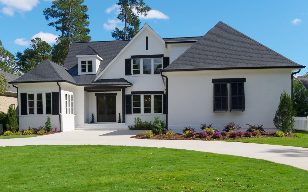 A white house with a black roof and a driveway. The house is large and has a lot of windows.