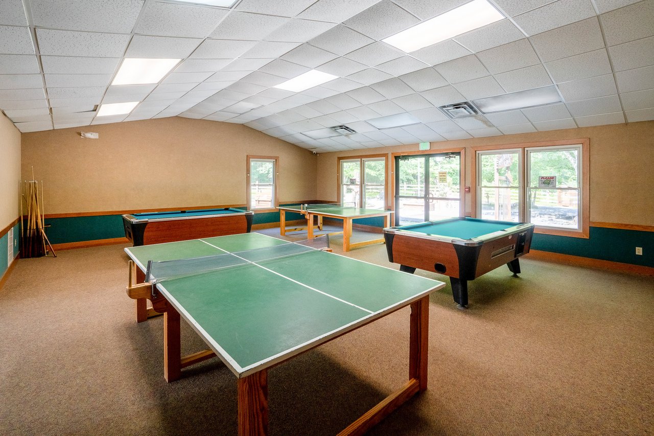 A recreational room features a ping pong table, pool table, and billiards table, with natural light streaming in from large windows on one side