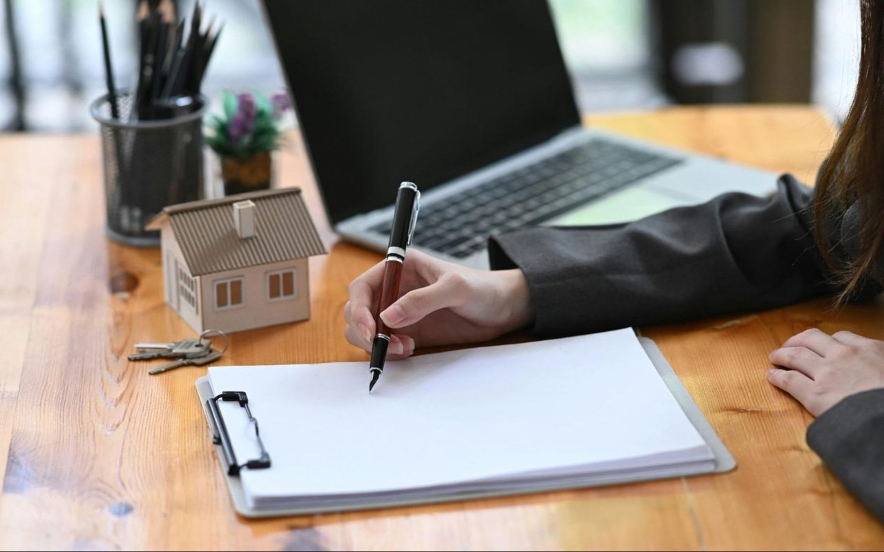  A person's hand, presumably that of a real estate agent or client, is seen signing a document on a clipboard with a pen. 