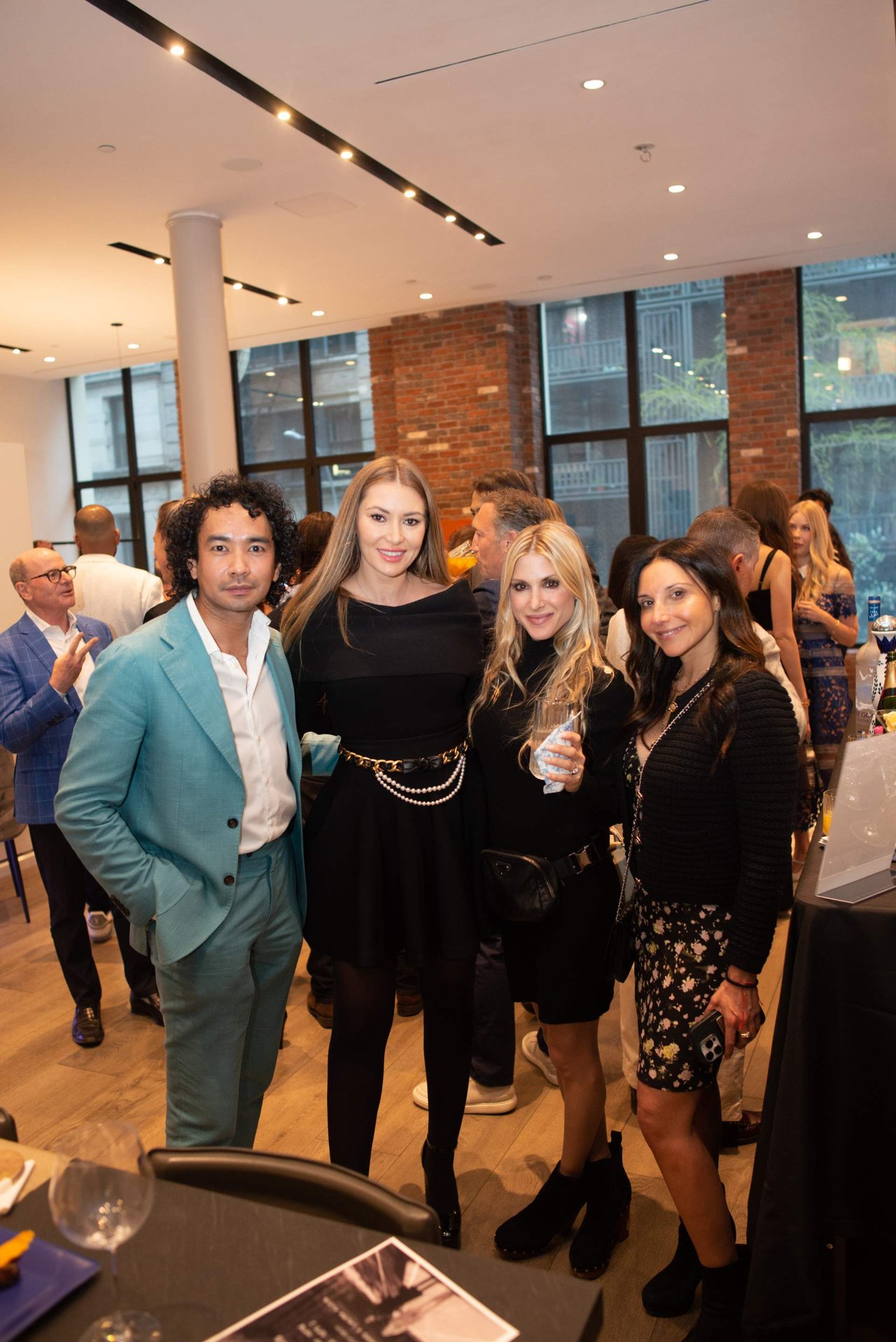 Four guests pose together at a crowded indoor event, with other attendees mingling behind them.