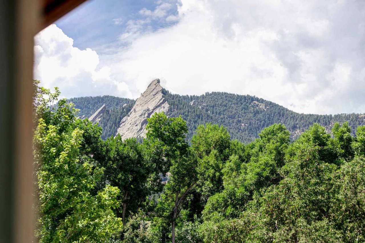 Flatirons mountain peak through summer trees, dramatic rock face against blue sky and white clouds.