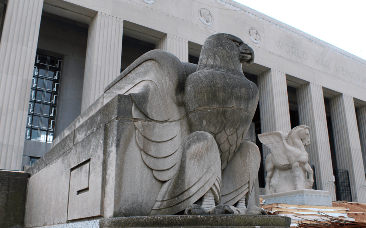  A large, weathered stone sculpture of an eagle with its wings partially spread sits prominently in front of a grand building with tall columns.