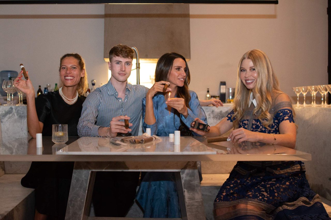 Four people gather around a marble table, smiling and holding cigars.