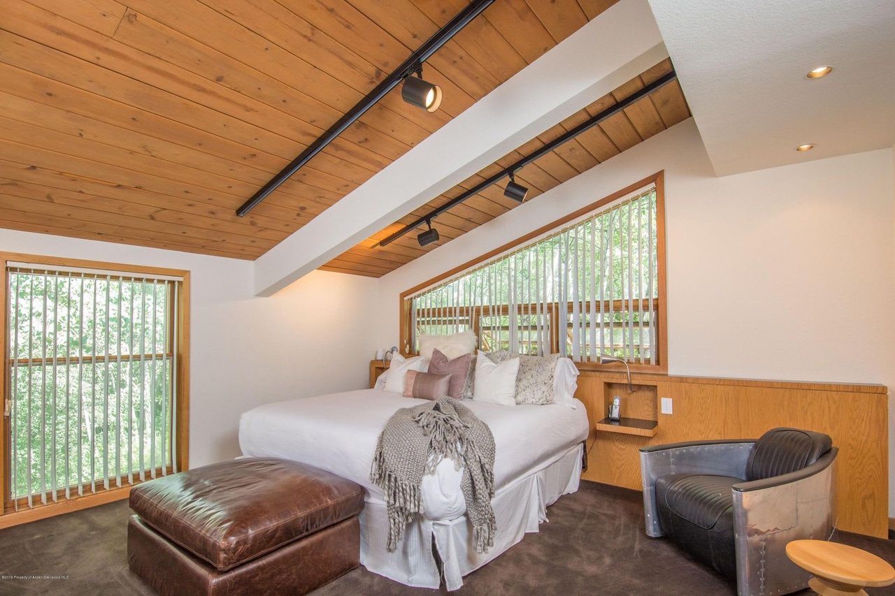 Bedroom with a wood-paneled vaulted ceiling, large bed, tall windows, and leather seating.