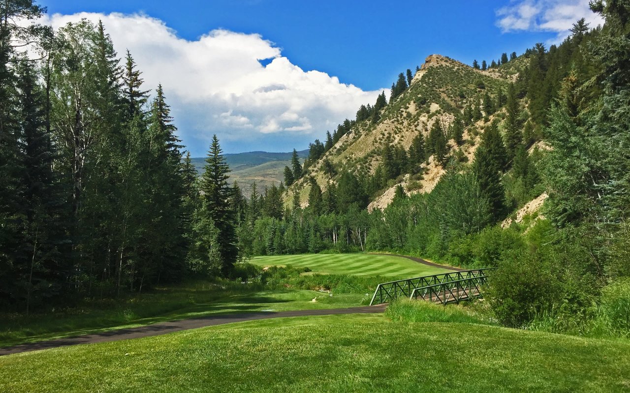 The Beaver Creek Golf Course in Colorado with a lush green fairway, quaint white bridge, and mountain backdrop.