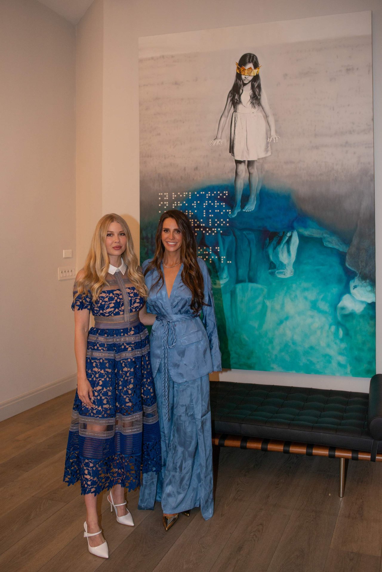 Two women in blue dresses pose beside a large artwork in an interior gallery.