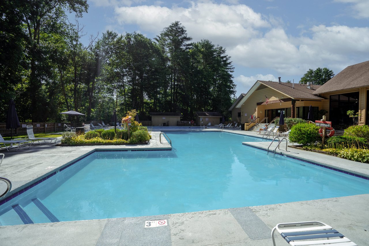 Alt=Outdoor swimming pool area surrounded by lounge chairs, with trees in the background and a partly cloudy sky.