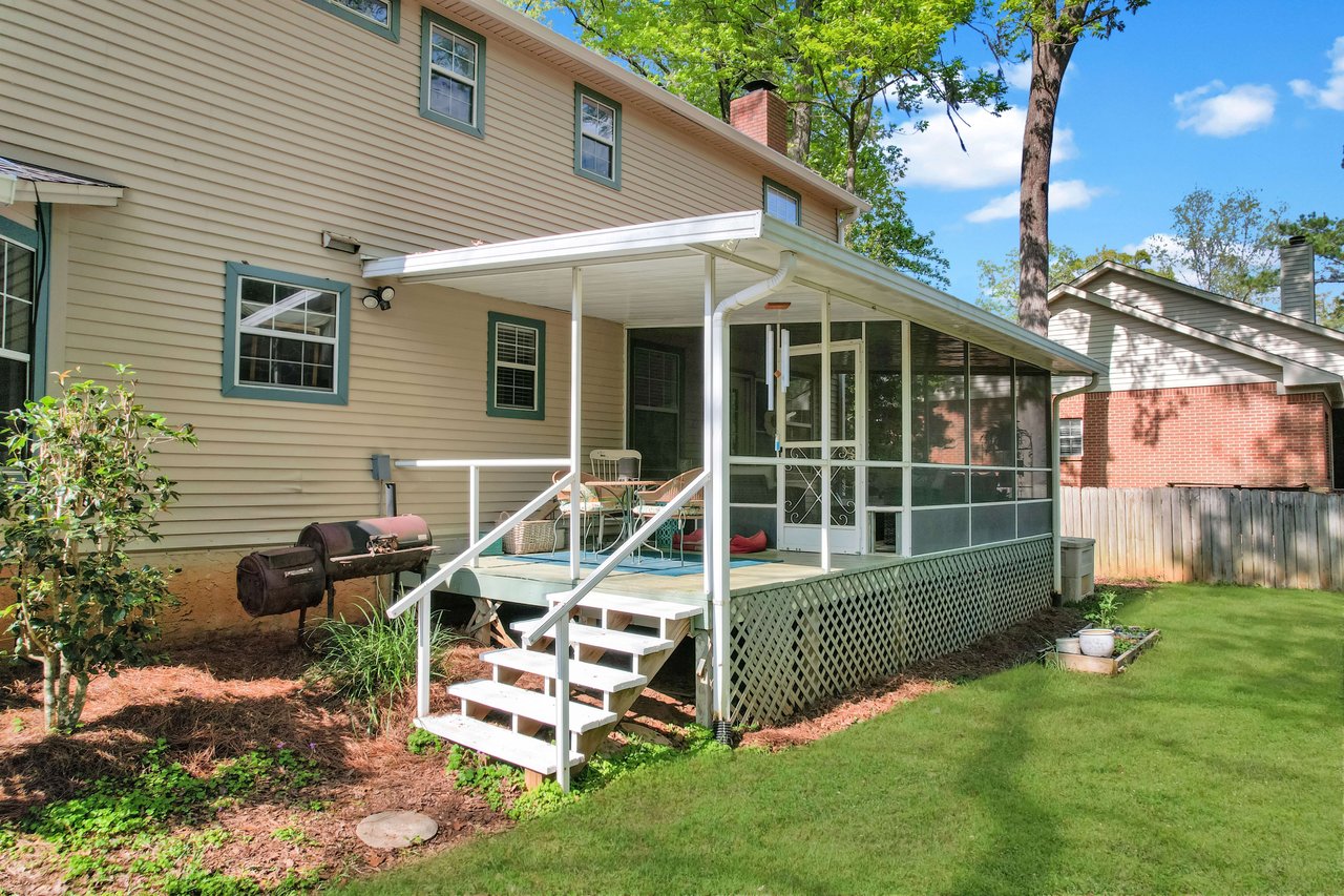 An exterior view of a two-story house with a well-maintained lawn. The house has a small back porch and is surrounded by greenery.