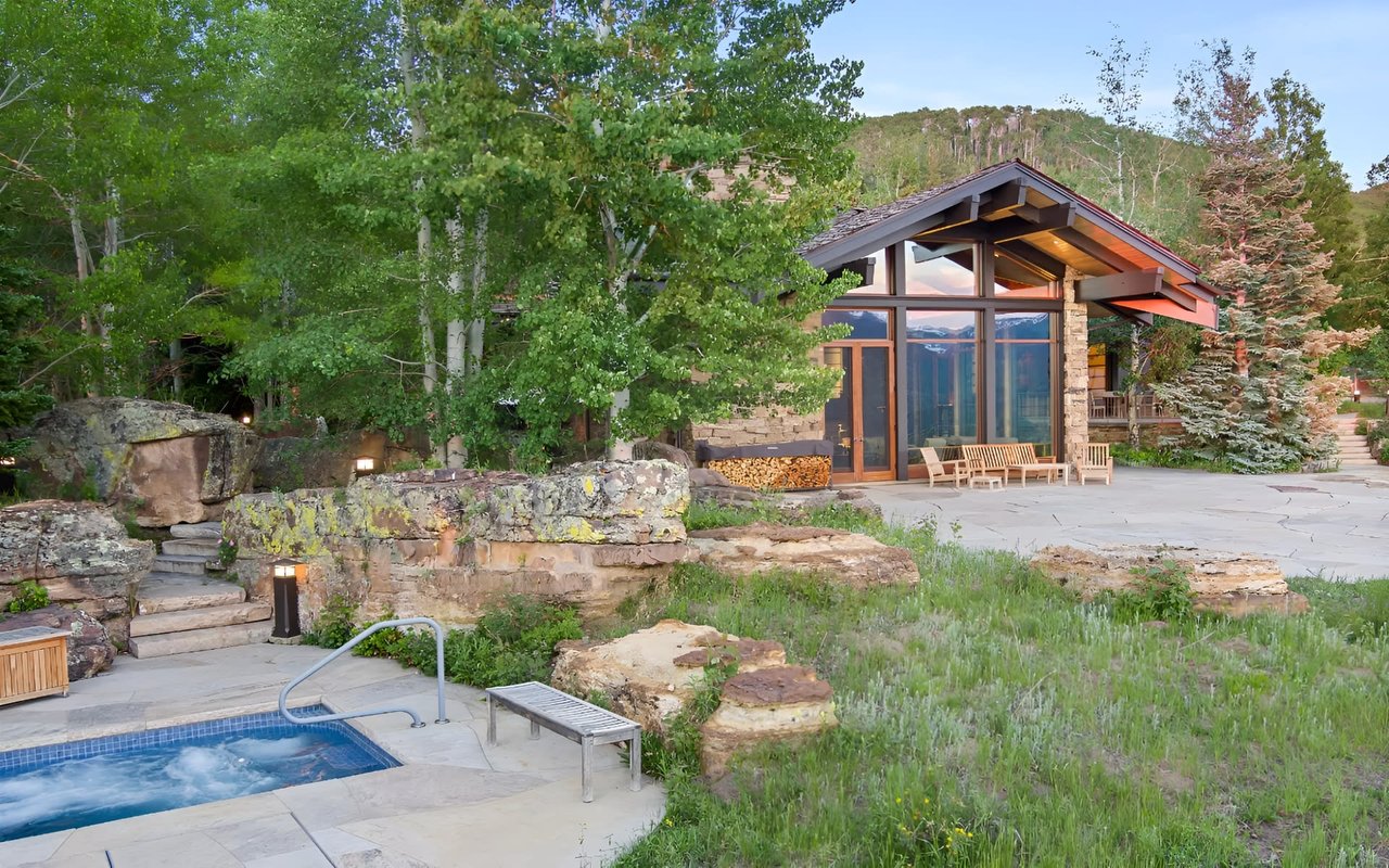 Modern stone-and-glass home beside a stone patio and pool, surrounded by trees and wooded hills.