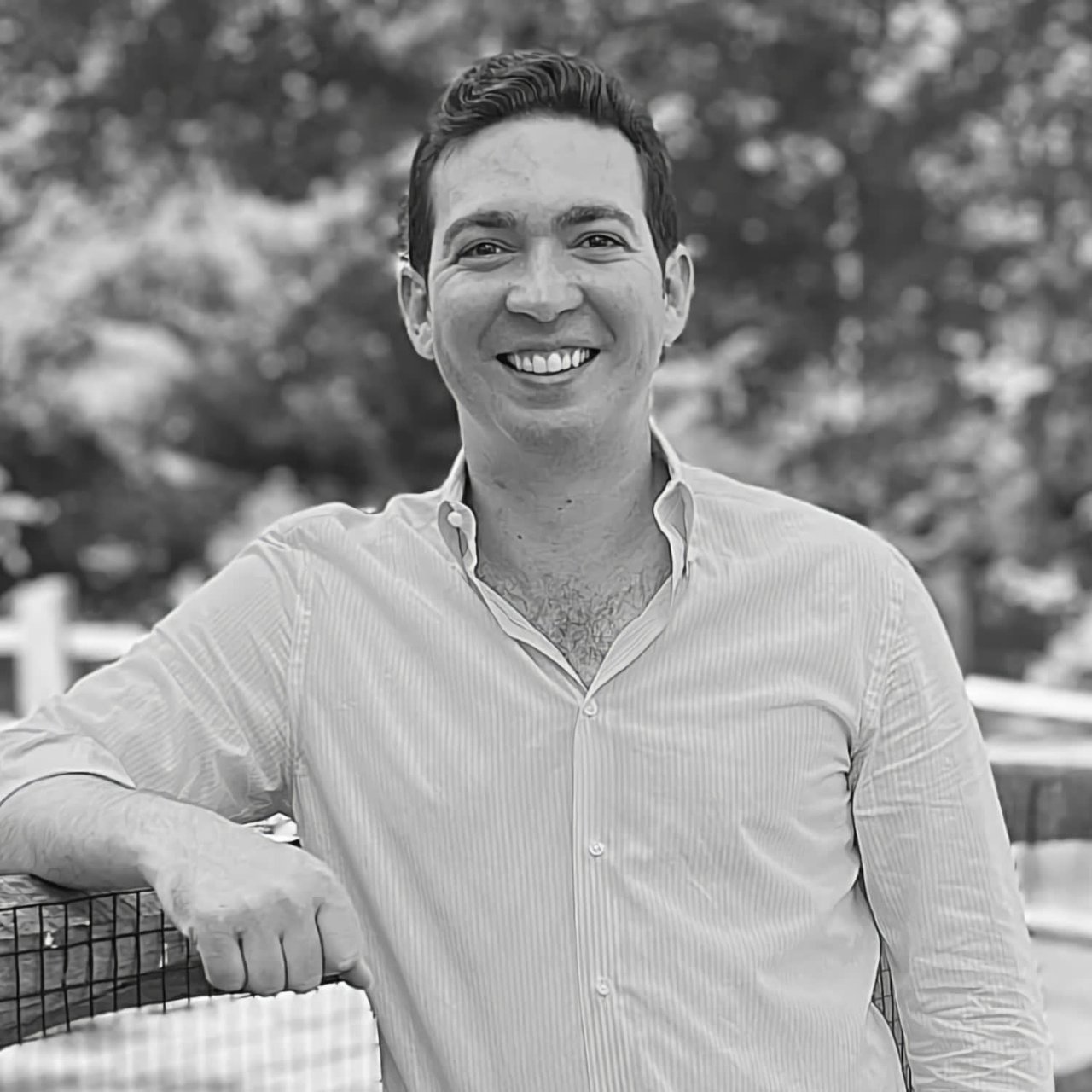 George D'Ambrosio in a button-up shirt smiles while leaning on a fence, with trees blurred in the background. The image is in black and white.