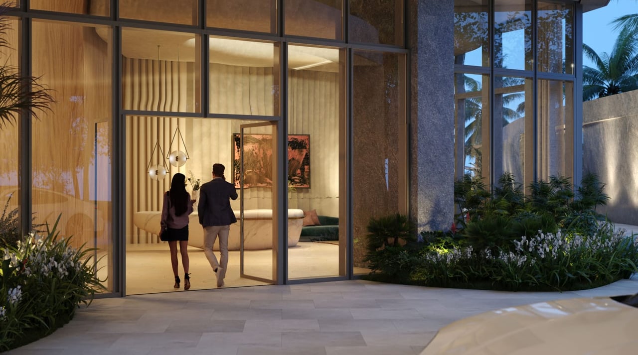 Glass-fronted residential entrance with landscaped beds and two people entering a warmly lit lobby.