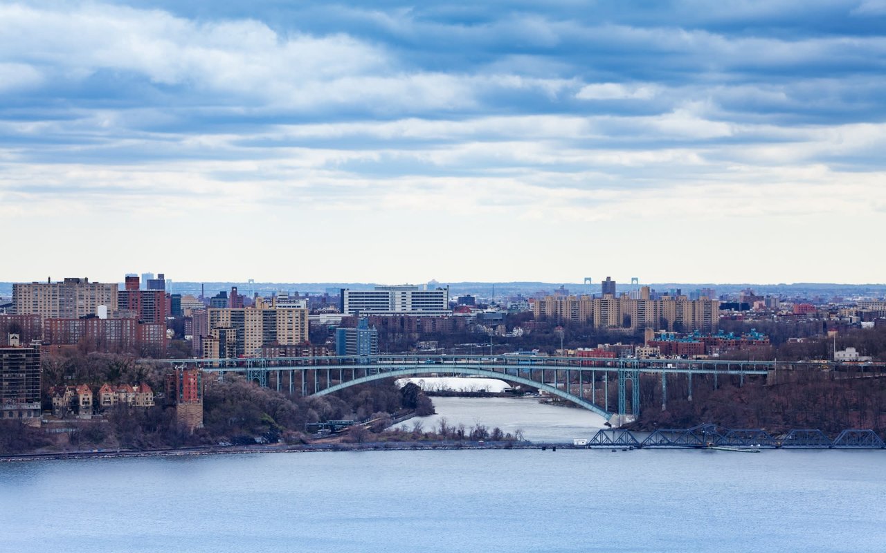 a steel suspension bridge spanning the Hudson River, the bridge is seen from the New Jersey side, with the towers in the foreground and the city skyline of Manhattan in the background.