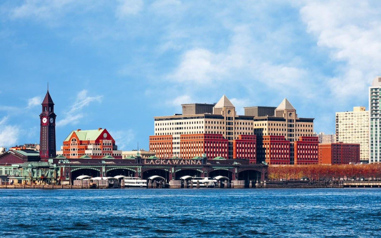 Hoboken, New Jersey's waterfront area with modern high-rises and older buildings, a clock tower, and a bridge over water.