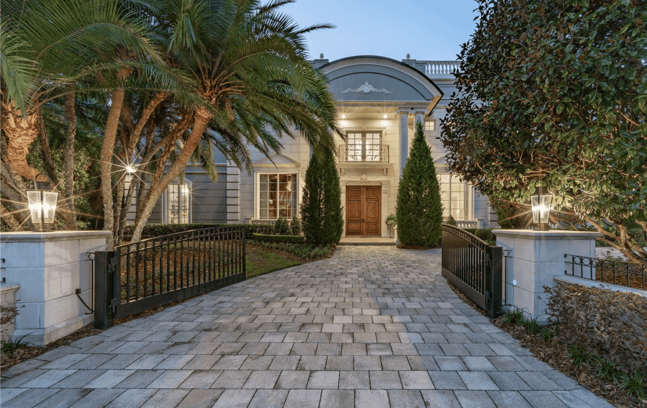 A two-story house surrounded by trees has a metal entrance gate, a driveway, and front lawns on either side.