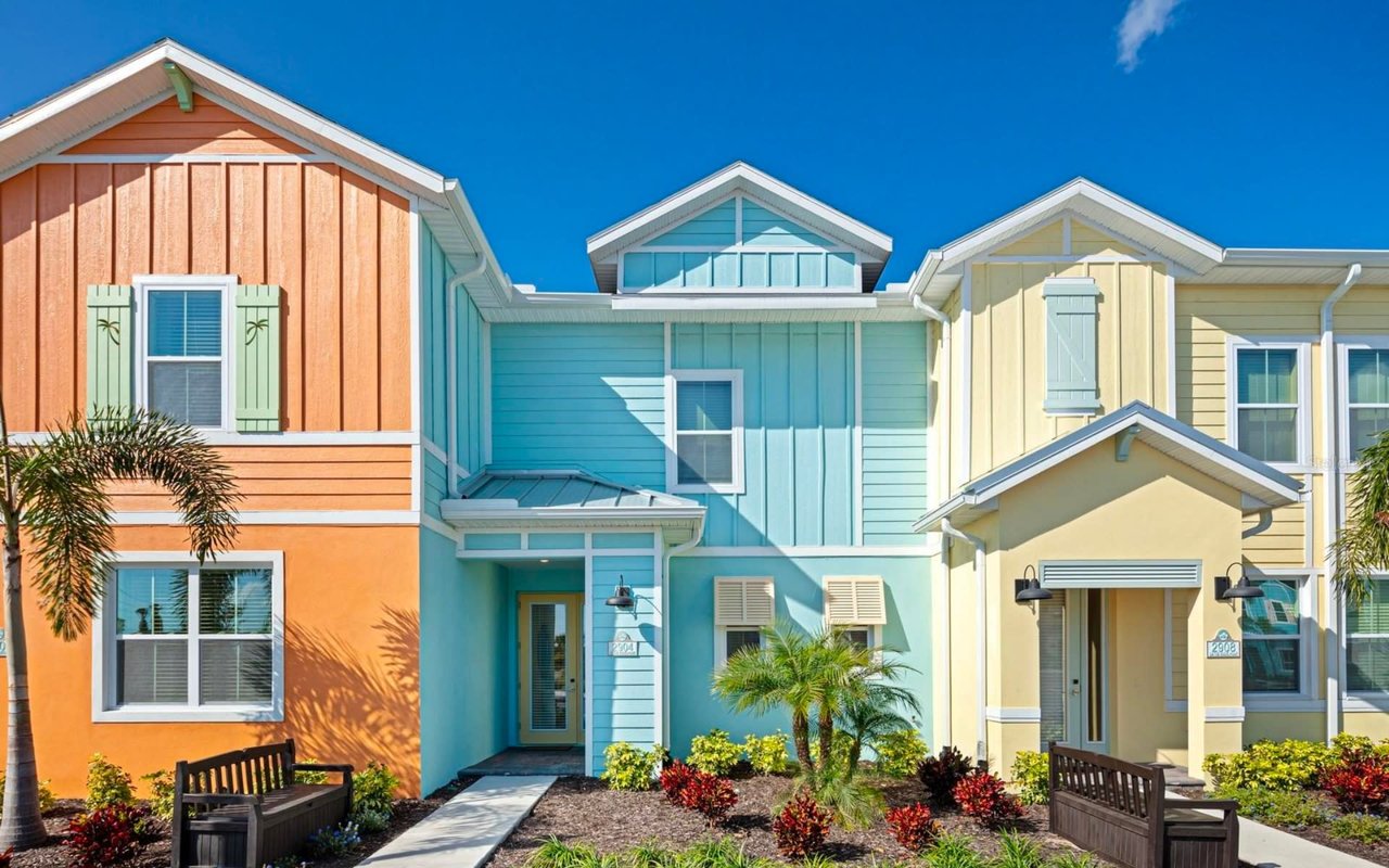A row of vibrant houses, each with a front garden and benches, bordered by palm trees, in a residential area.