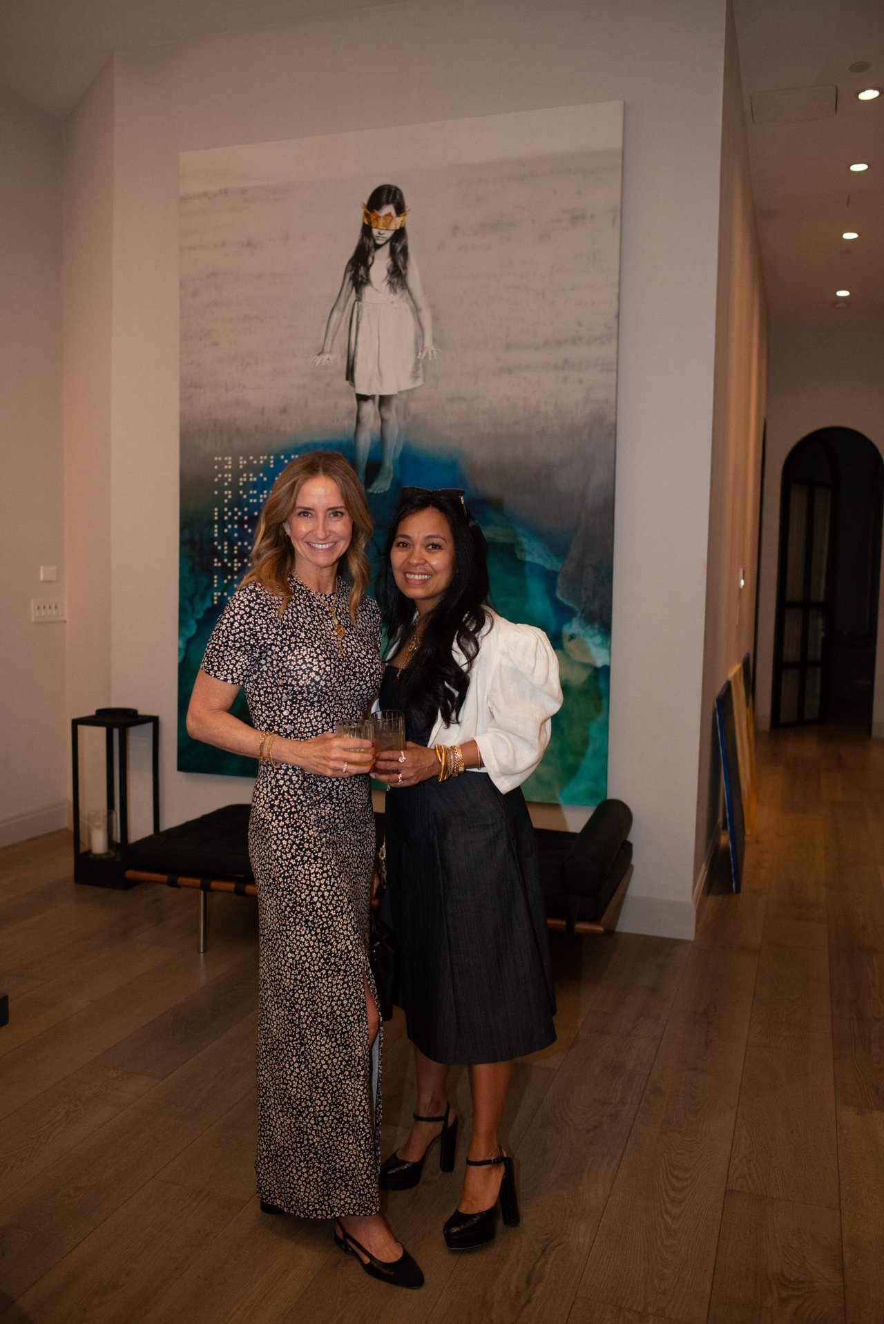 Two women pose with drinks in front of a large artwork.
