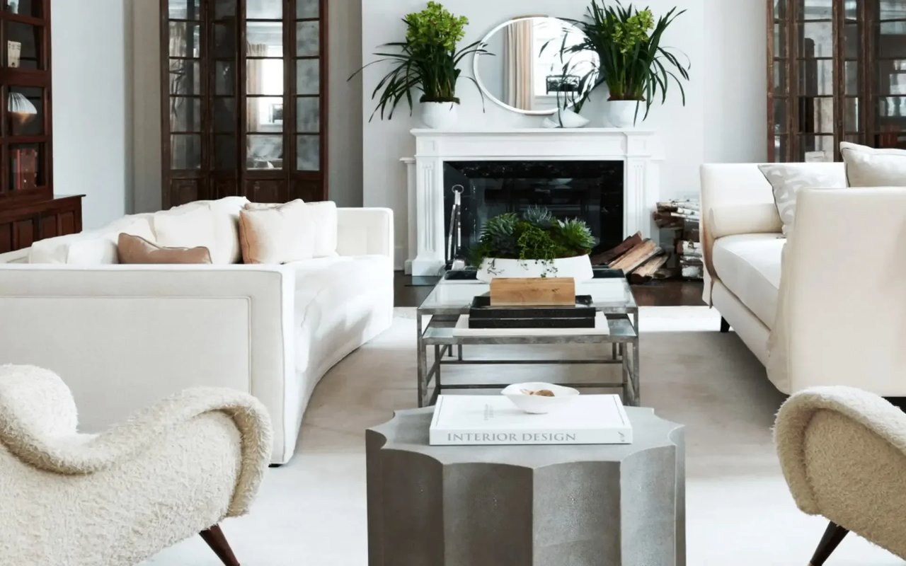 A modern living room with white sofas, a glass coffee table, and a marble fireplace, with plants, books, and a side table.