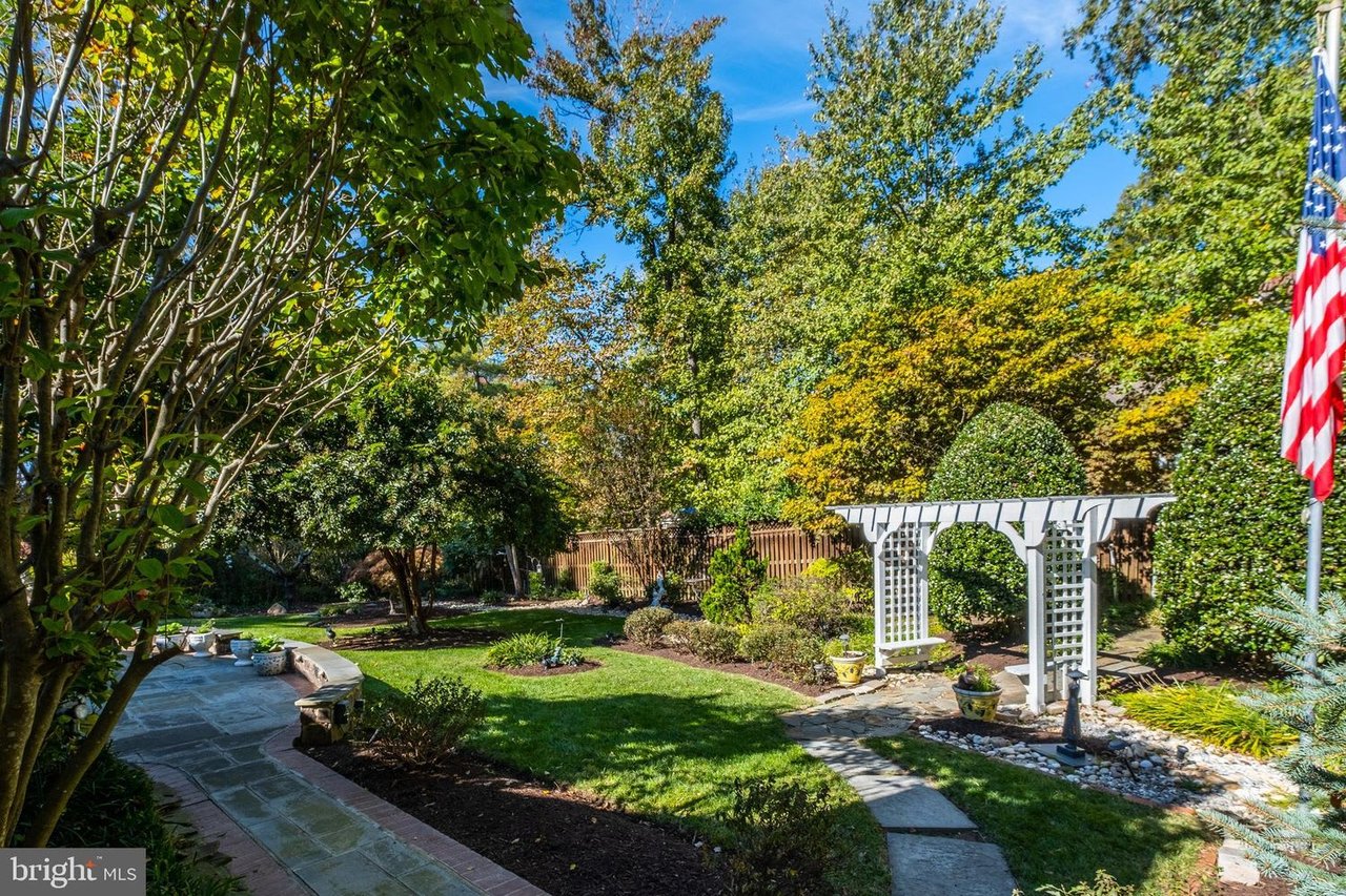 Landscaped backyard with mature trees, a stone patio, winding path, and white pergola.