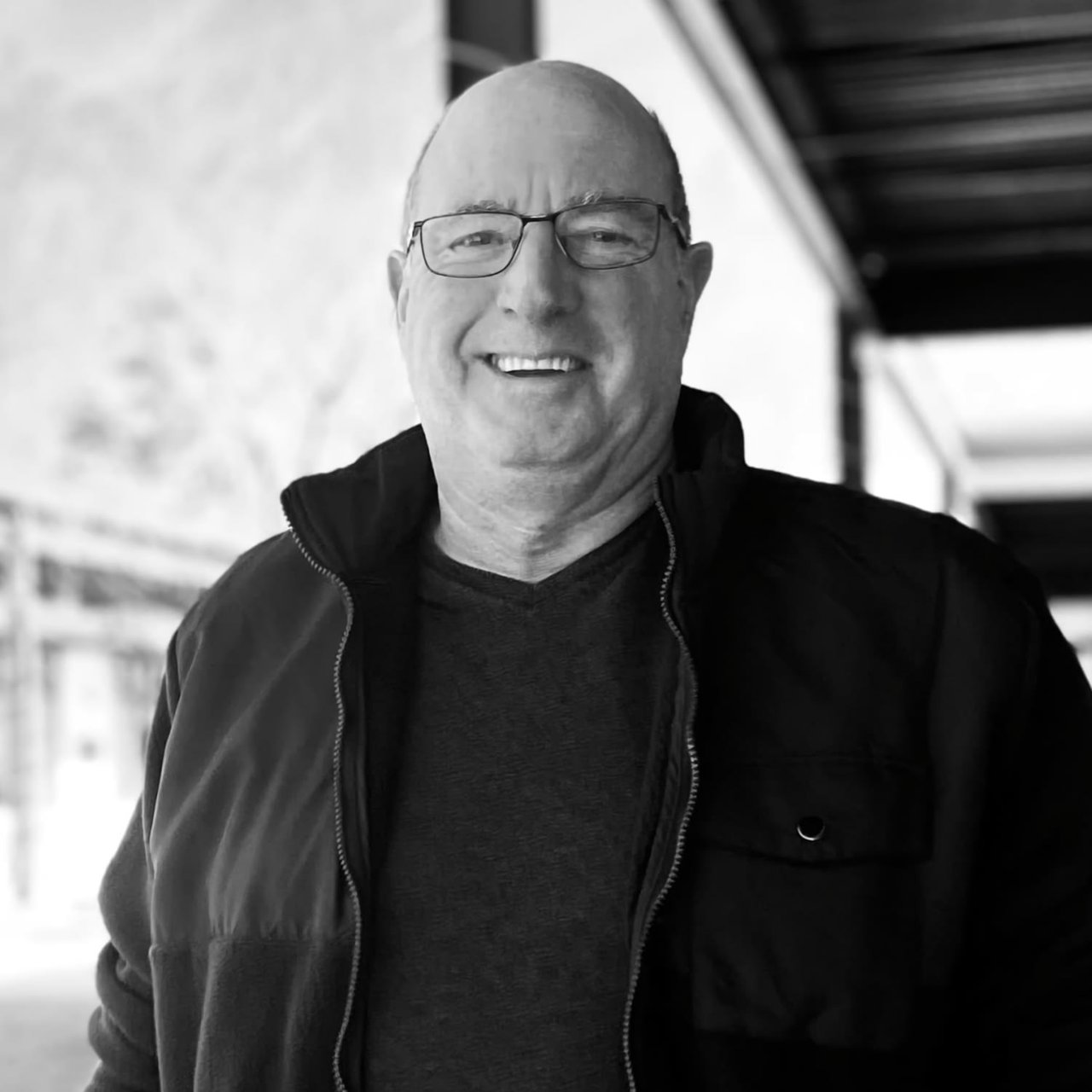 Peter Michaelis wearing glasses and a zip-up jacket smiles while standing outdoors under a covered walkway. The image is in black and white.