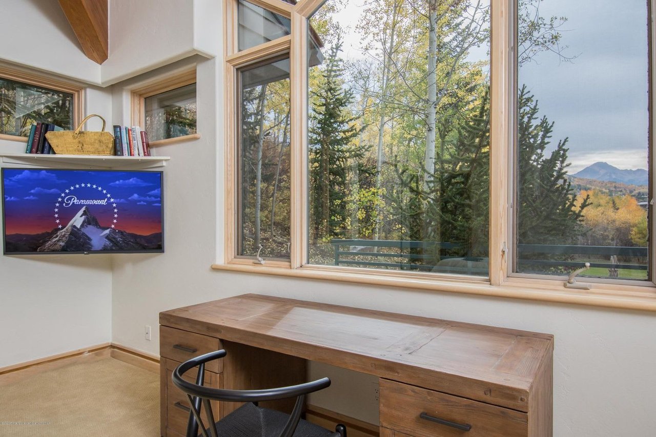 Home office with a wooden desk beside large windows overlooking autumn trees and distant mountains.