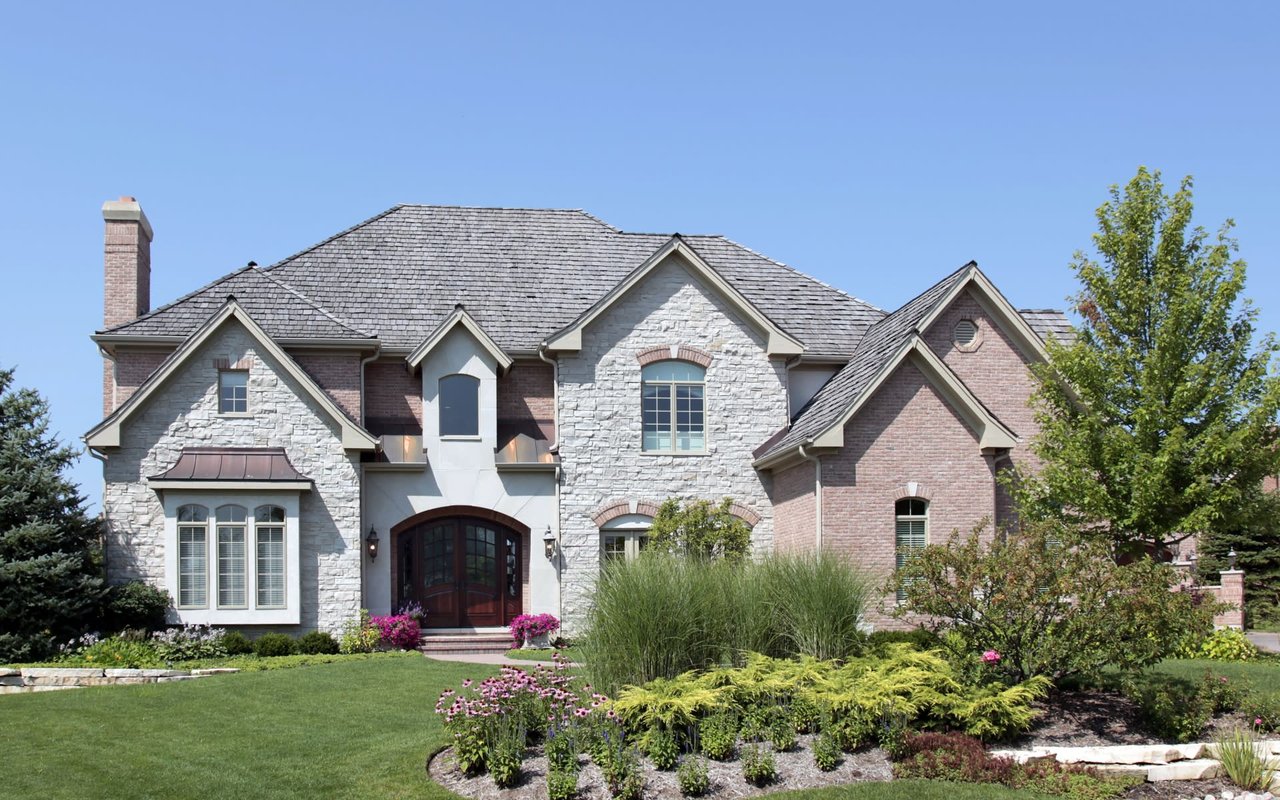 A large brick and stone house with a slate roof sits amidst a spacious lawn with mature trees.