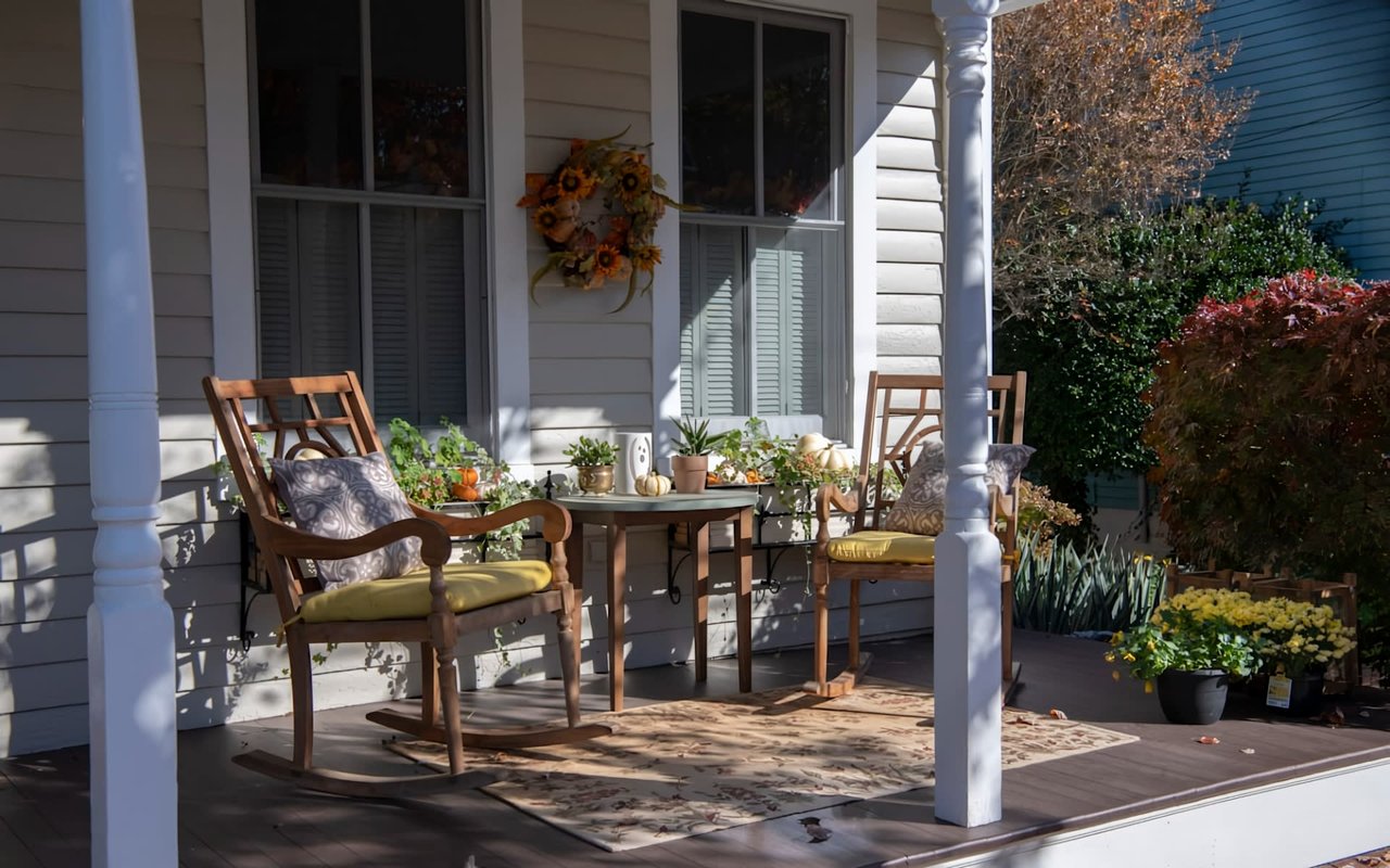  A front porch of a house decorated for autumn, featuring two wooden rocking chairs with yellow cushions and patterned pillows.