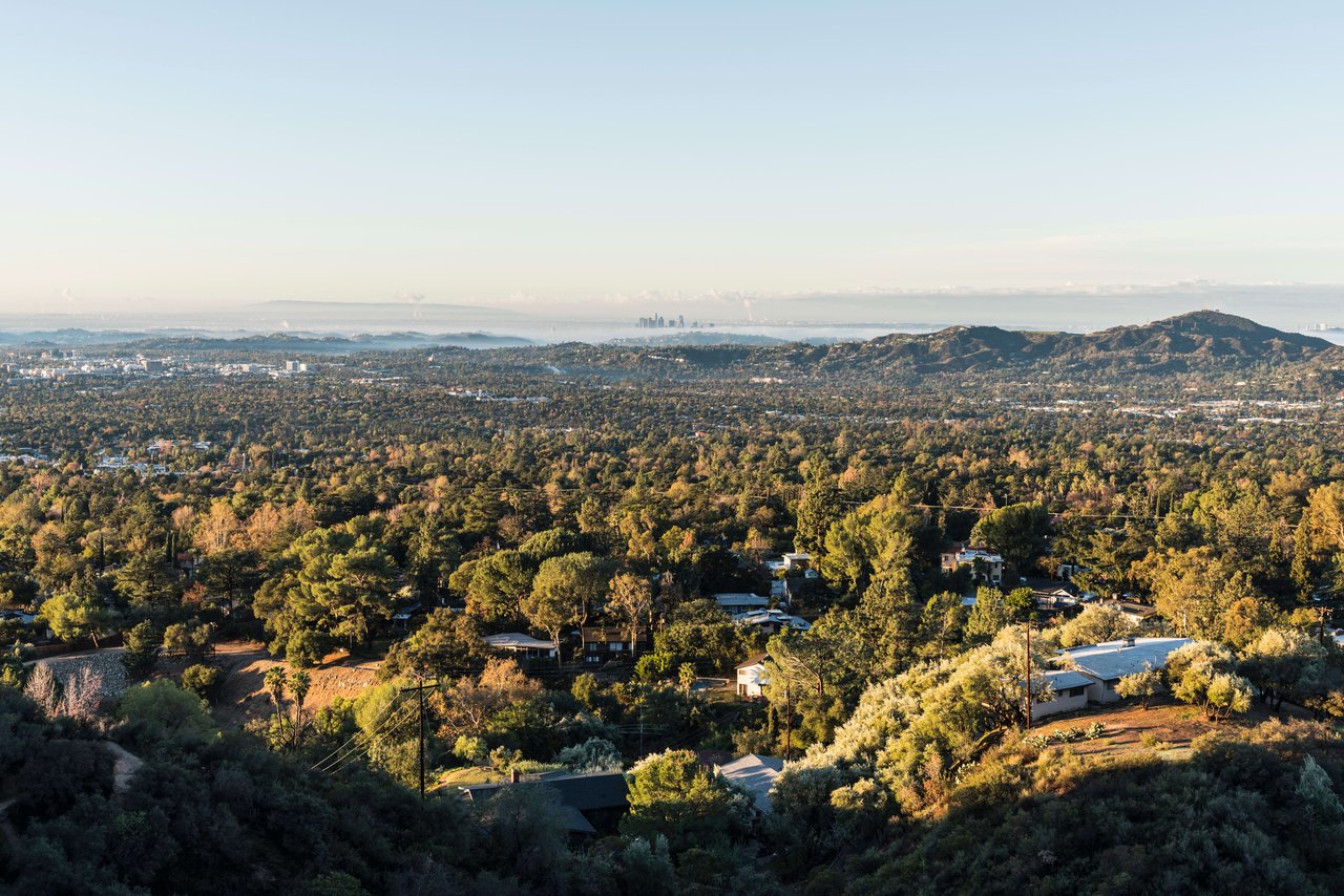 A view of a city from a hilltop. The city is spread out below, with a variety of buildings, trees, and streets.