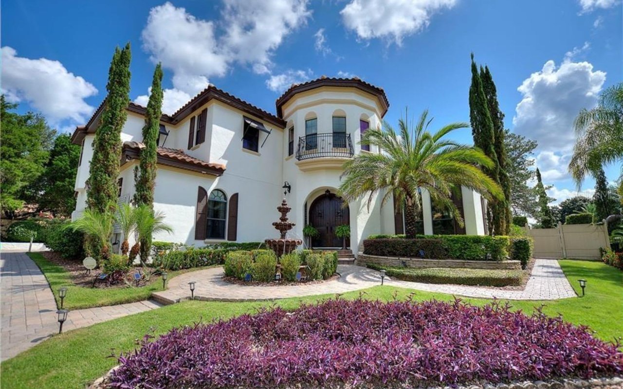 A grand Georgian-style house with a fountain, a balcony, a front lawn, and a curved driveway lined with trees.