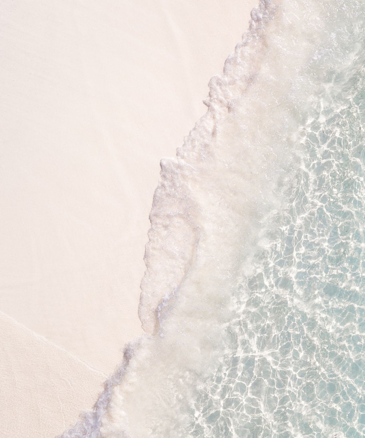  An aerial shot captures the serene meeting of a pristine white sandy beach and crystal-clear, pale turquoise ocean water.